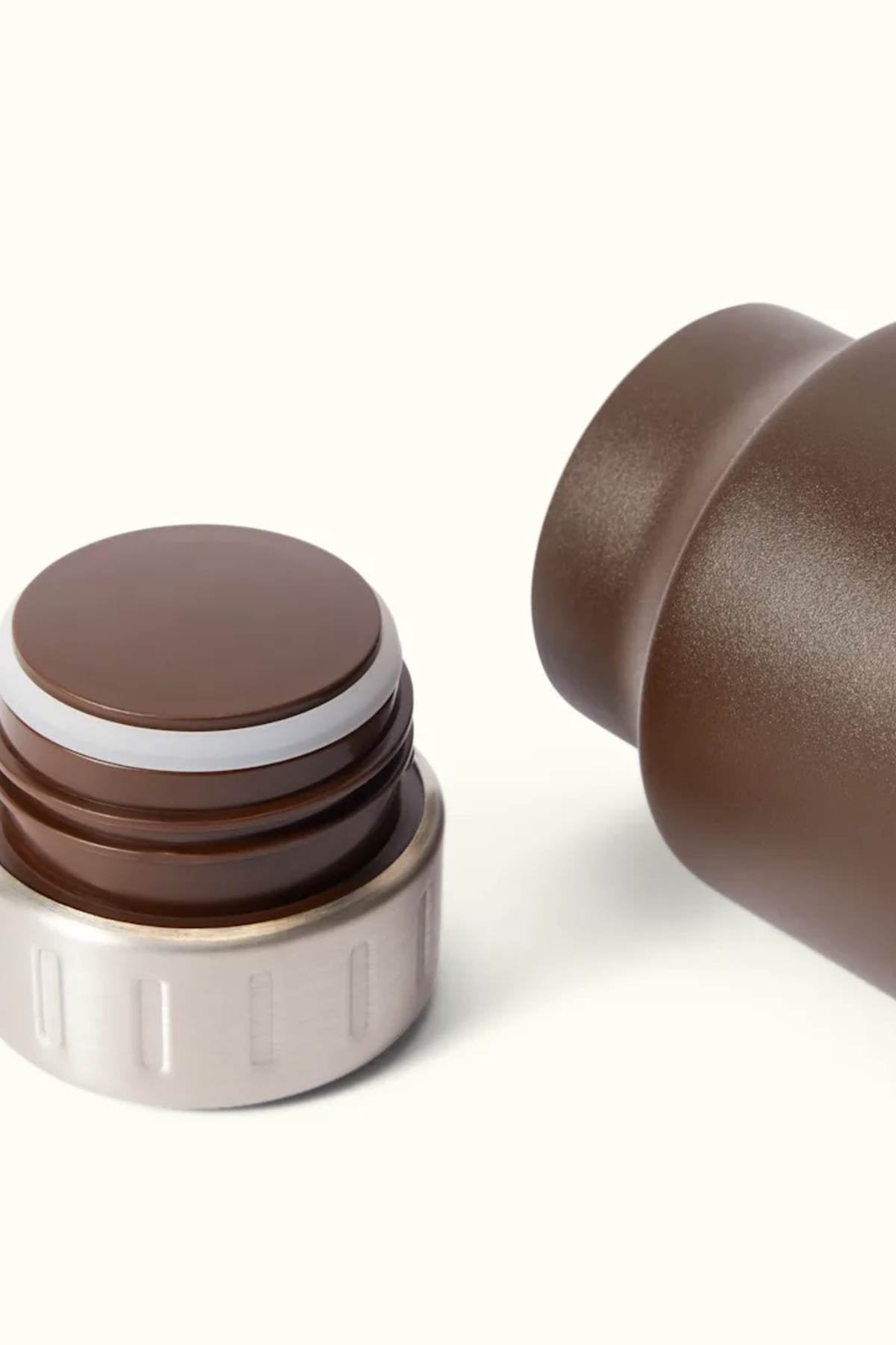Close-up of the RM Williams Water Bottle, a brown, vacuum-insulated travel mug with a detachable lid. The lid is displayed on the left, highlighting a metallic ring and a secure seal. The BPA-free bottle's opening is visible on the right against a light background.