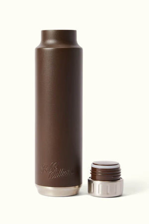 A stainless steel, brown water bottle from RM Williams has its 'R.M. Williams' logo embossed near the bottom, with the cap removed. Its vacuum-insulated design maintains long-lasting temperature control. The white cap with brown accents is positioned nearby on an off-white background, ensuring a BPA-free experience.