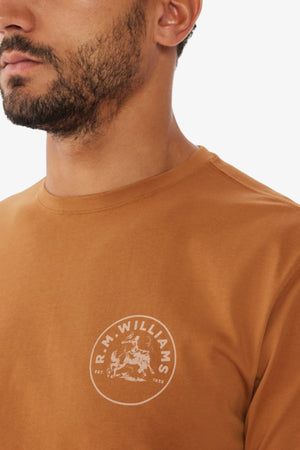 A man wears a brown RM Williams Wondai T-Shirt with the logo on the chest, crafted from lightweight cotton jersey. The graphic print highlights the brand name and design, while his partially visible face draws attention to the shirt's details.
