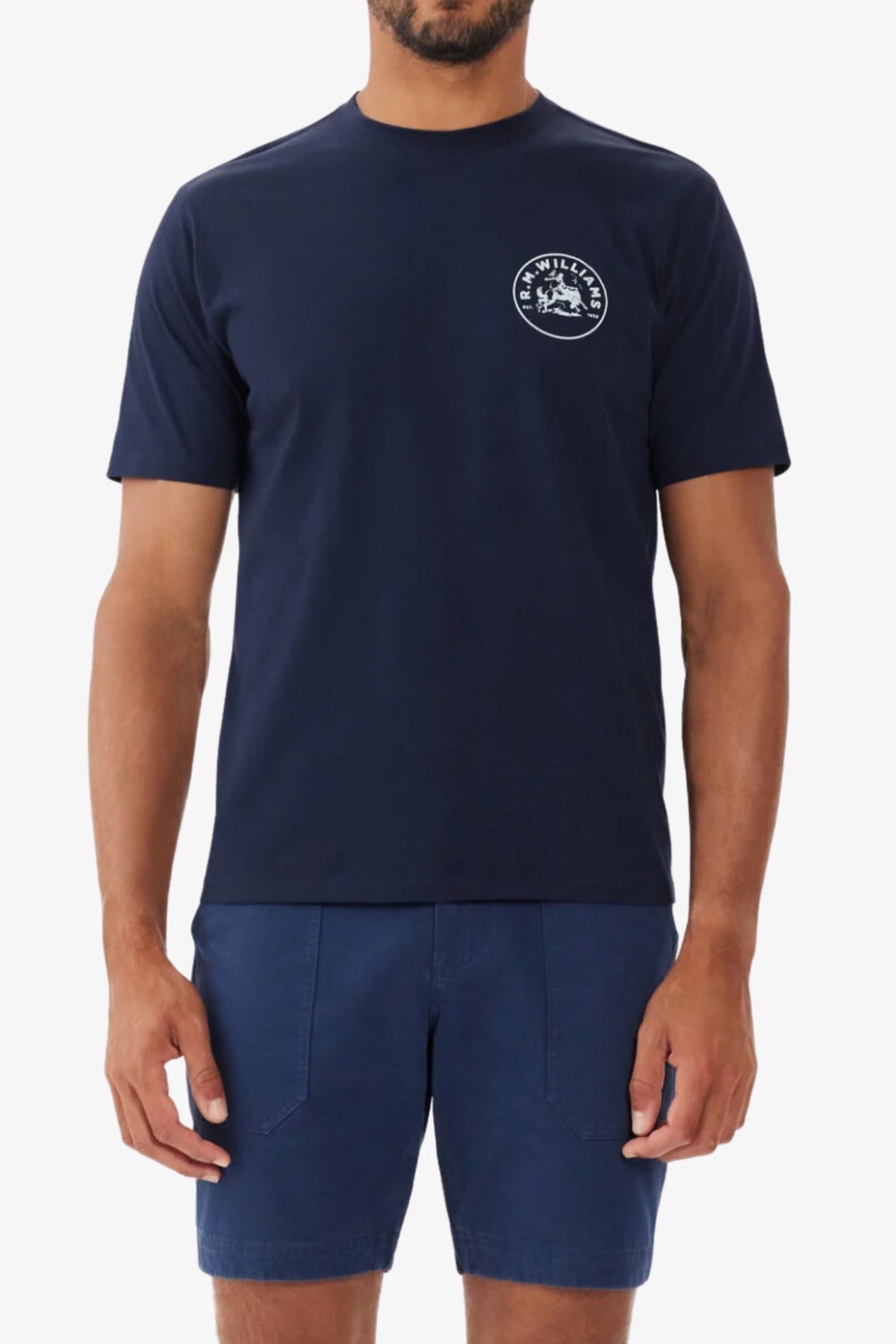 A man wears a navy RM Williams Wondai T-Shirt, made from lightweight cotton jersey, featuring a small white circular logo on the left chest, paired with matching navy shorts, standing against a plain white background.