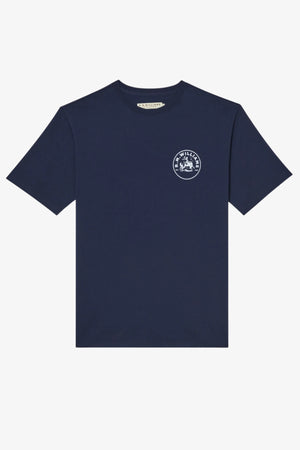 The RM Williams Wondai T-Shirt is a plain navy blue, short-sleeved cotton jersey with a small white archive-inspired circular graphic and text on the left chest. It is shown laid flat on a white background.