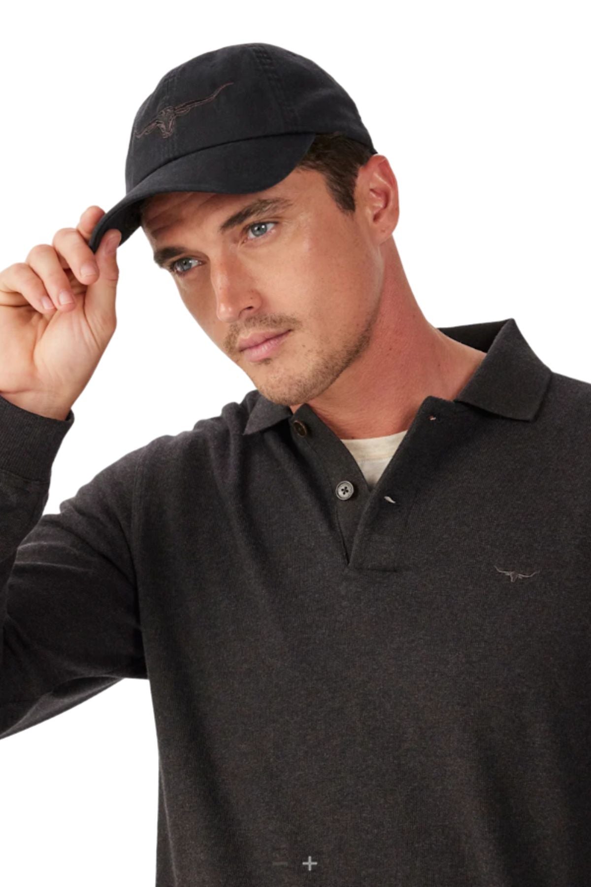 A man wearing the RM Williams Longhorn Twill Cap—dark organic cotton with an embroidered Longhorn—looks to the side as he adjusts the cap’s strap. He pairs it with a dark collared shirt against a white background.