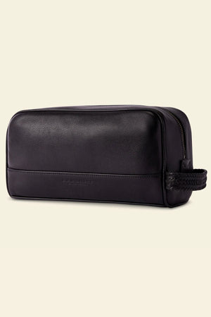 The RM Williams Farrier Signature Washbag is a black tumbled grain leather toiletry bag with a rectangular shape, zippered top, and braided end handle, shown against a plain light background.