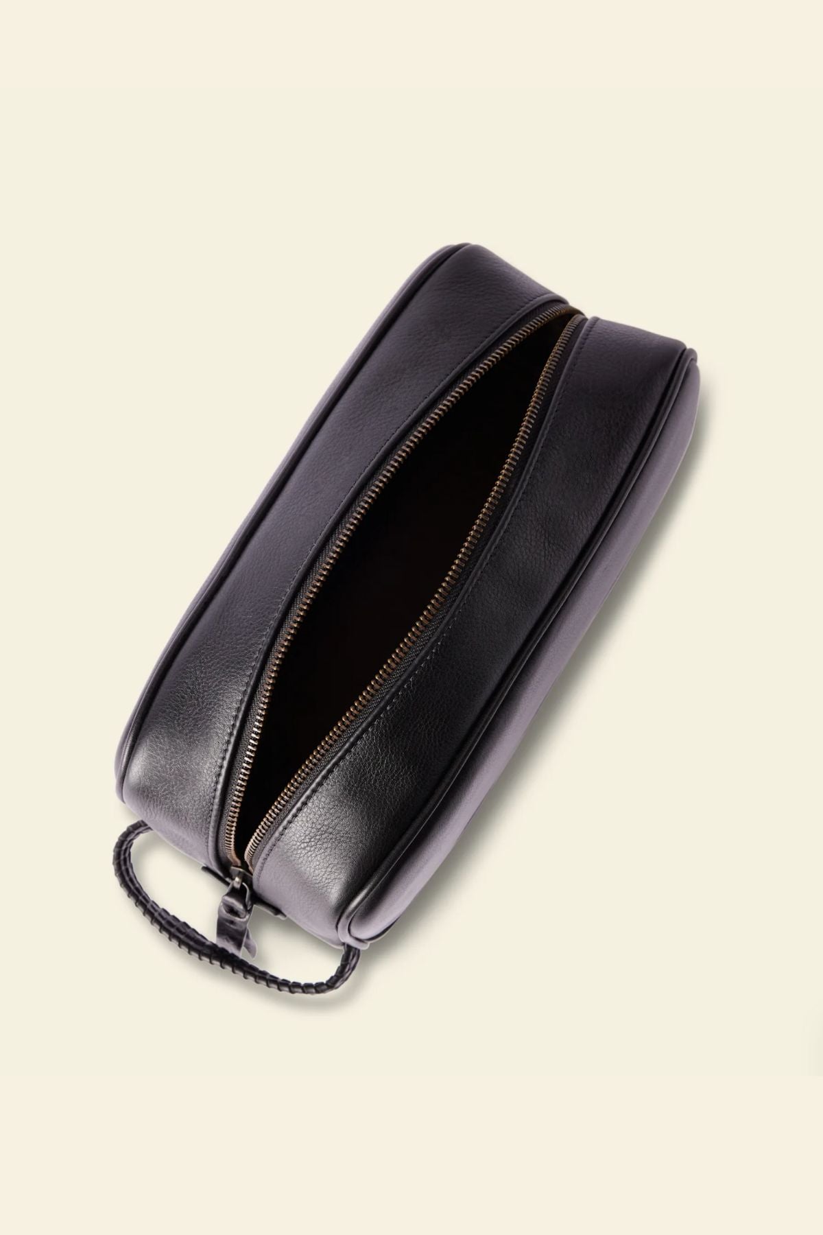 The RM Williams Farrier Signature Washbag, crafted from black tumbled grain leather, features a textured handle, water-resistant lining, and a zippered top. Shown against a light beige background with its empty interior visible.