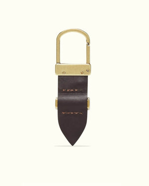 The RM Williams Drover Key Fob is a gold carabiner-style keychain with a dark brown, double butt leather design and two stitches on its pointed end. Antique brass hardware elegantly joins the metal and leather components against a plain light background.