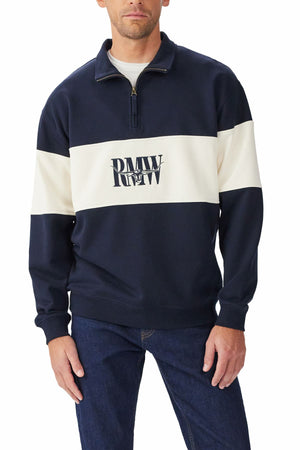 A man wears the RM Williams Heritage 1/4 Zip Sweatshirt in navy with a wide white stripe and "PMW RW" logo on the front, paired with dark blue jeans.