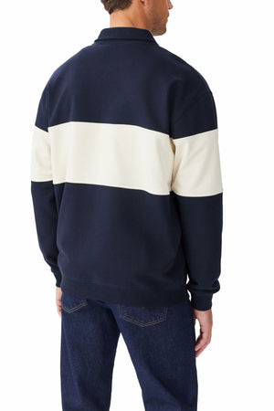 A man stands facing away, wearing the RM Williams Heritage 1/4 Zip Sweatshirt in navy with a wide white horizontal stripe across the back and sleeves, paired with dark blue jeans.