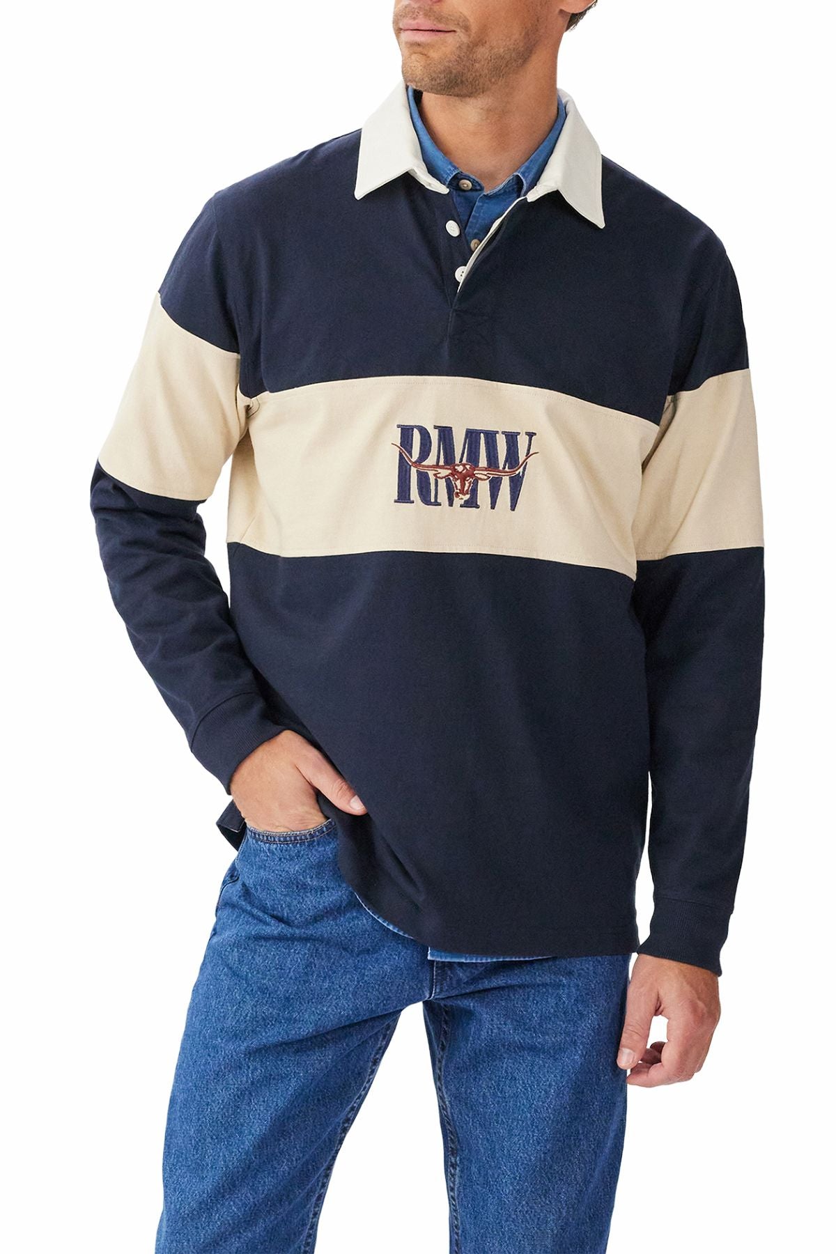 A man wears the RM Williams Heritage Rugby in navy blue and beige brushed organic cotton with a "PMW" logo across the chest, paired with blue jeans. He stands with one hand in his pocket, looking slightly to the side.