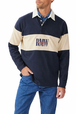 A man wears the RM Williams Heritage Rugby in navy blue and beige brushed organic cotton with a "PMW" logo across the chest, paired with blue jeans. He stands with one hand in his pocket, looking slightly to the side.