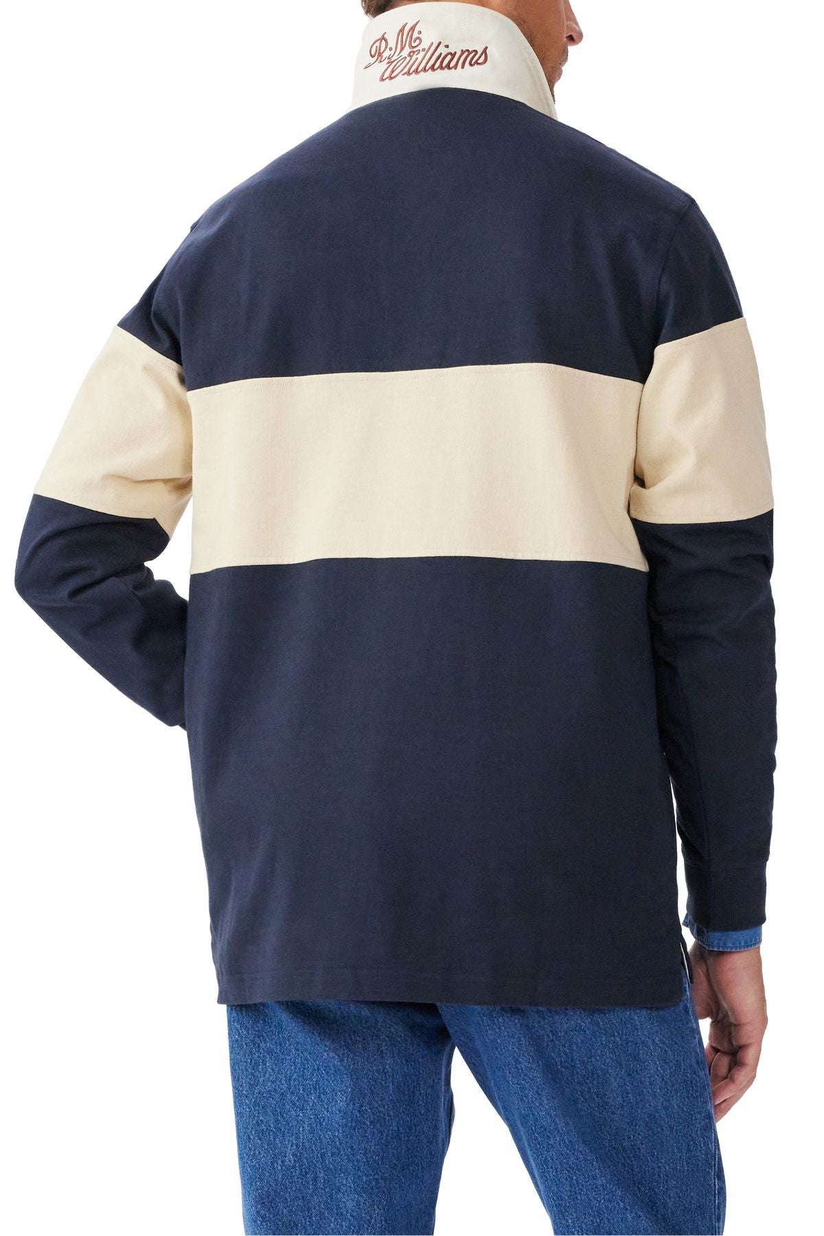 A person is seen from behind wearing the RM Williams Heritage Rugby jacket in navy blue and beige with "RL McWilliams" embroidered on the collar, styled like a classic rugby shirt, and paired with blue jeans.