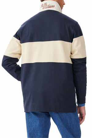 A person is seen from behind wearing the RM Williams Heritage Rugby jacket in navy blue and beige with "RL McWilliams" embroidered on the collar, styled like a classic rugby shirt, and paired with blue jeans.