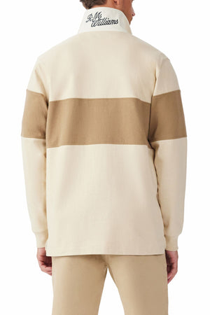 A person stands with their back to the camera, wearing the RM Williams Heritage Rugby—a beige heavyweight brushed cotton long-sleeve shirt featuring a broad brown stripe across the upper back and arms, plus "R.M. Williams" embroidered on the collar.