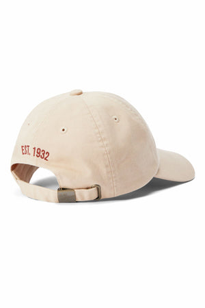 The RM Williams Heritage Twill Cap in beige organic cotton is shown from the back, featuring an embroidered heritage logo, red "EST. 1932" on the left, a metal clasp adjustable strap, and a curved brim.