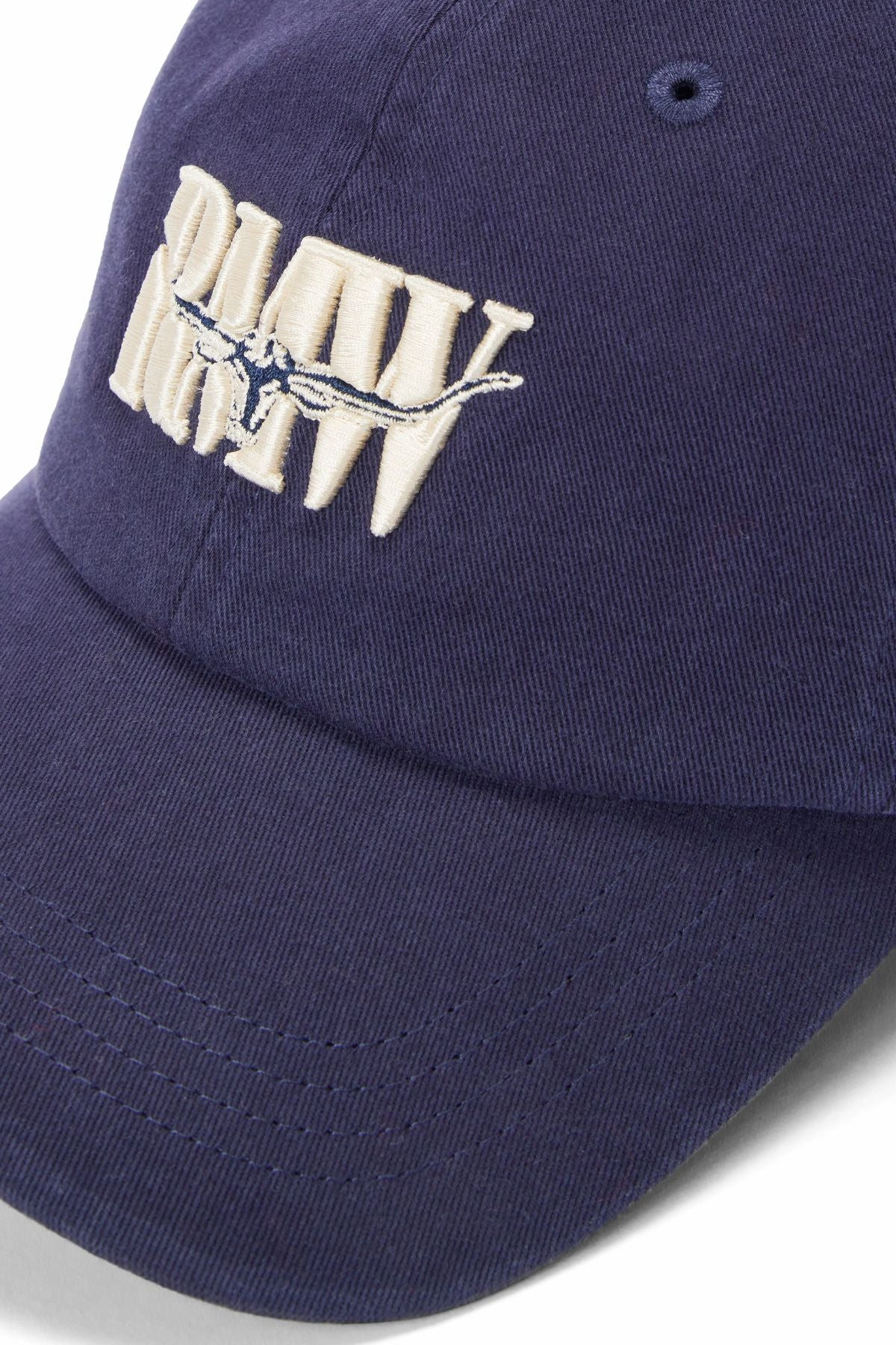 Close-up of the RM Williams Heritage Twill Cap in navy blue organic cotton, featuring "RAW CREW" embroidered in white with a barbed wire graphic through the text.