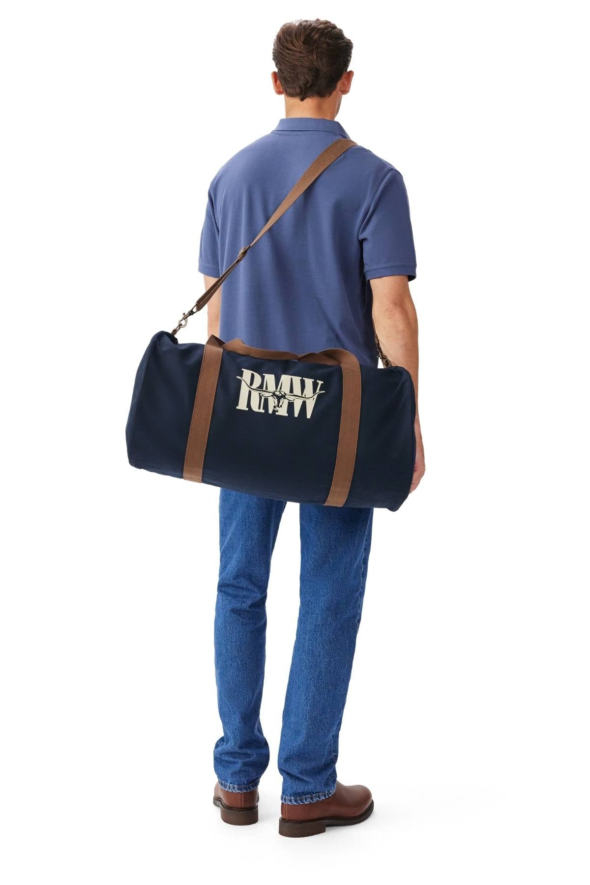 A man in a blue polo, jeans, and brown shoes stands facing away, carrying the RM Williams Heritage Bag—navy cotton canvas with an adjustable crossbody strap and white heritage logo—over his shoulder.