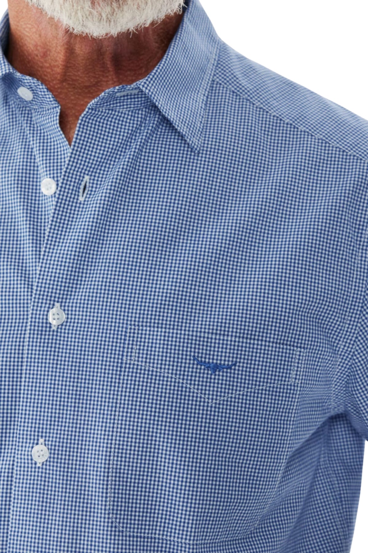 A close-up reveals part of an older man's chin and neck clad in a blue and white checkered RM Williams Hervey Shirt, made from breathable cotton. The shirt highlights a button-down collar, a front pocket, and buttons down the front. The man has a white beard and slightly wrinkled skin.