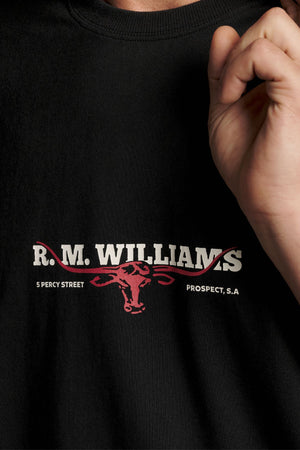 A person is wearing the RM Williams Holt Tee, a black relaxed fit shirt featuring βR.M. Williamsβ and a red bull logo, along with β5 Percy Streetβ and βProspect, S.A.β printed on it. The teeβs ribbed crew neck and premium Australian cotton enhance its comfortable style. A hand is partially visible near the shirt.