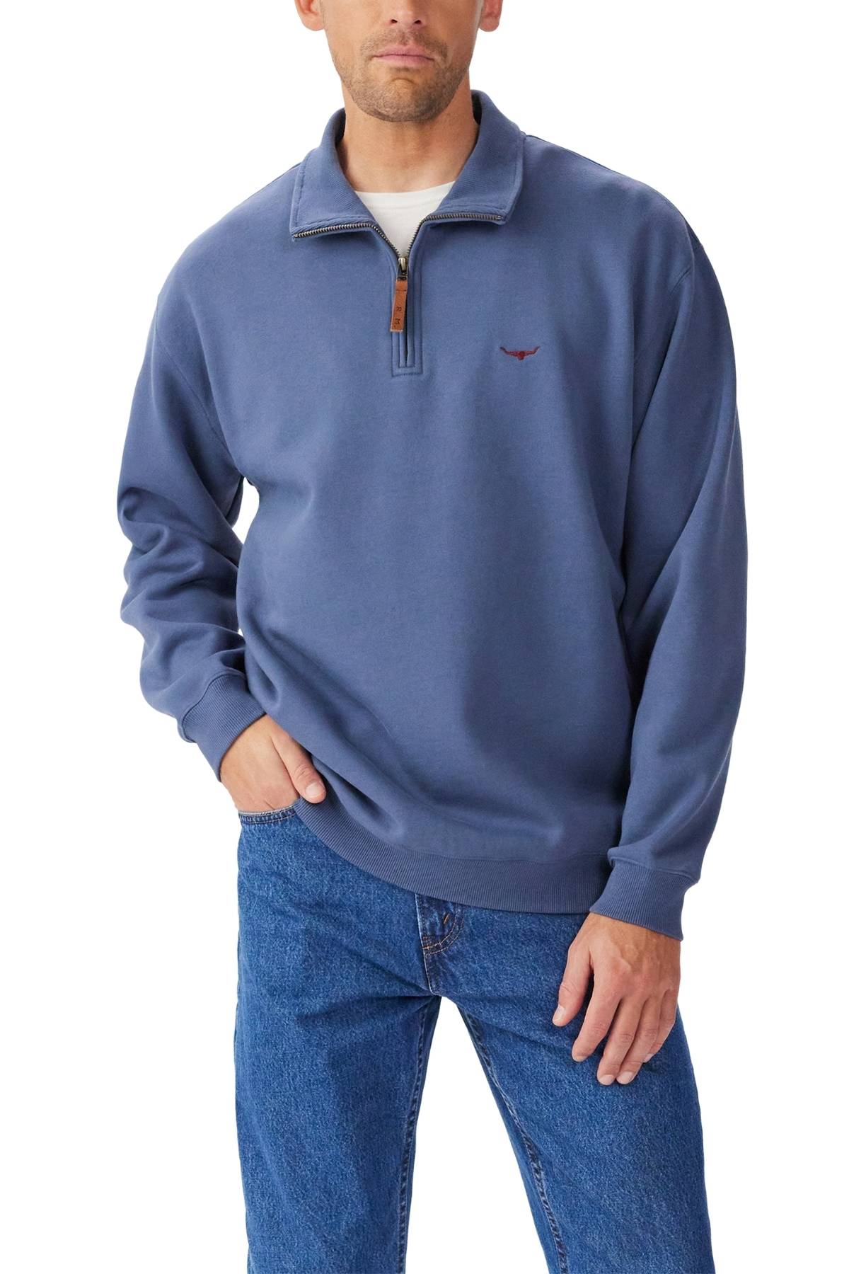 A man in the RM Williams Mulyungarie 1/4 Zip Sweatshirt—blue, organic cotton, small chest logo—and blue jeans stands with one hand in his pocket against a white background.