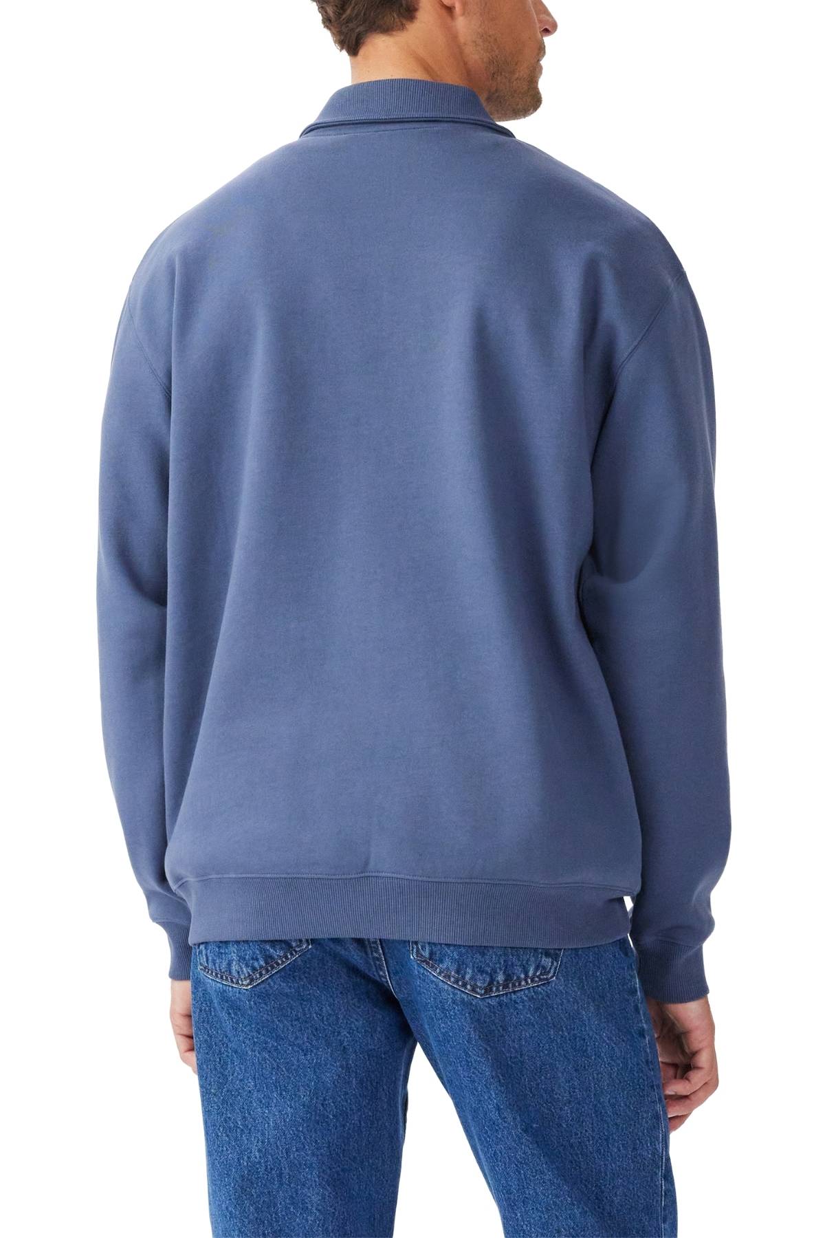 A man is seen from the back wearing the RM Williams Mulyungarie 1/4 Zip Sweatshirt in blue with relaxed fit, long sleeves, and ribbed cuffs and hem, paired with blue jeans. The organic cotton sweatshirt is set against a white background.
