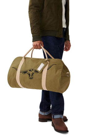 A person wearing a green jacket, blue jeans, and brown boots carries the RM Williams Nanga Canvas Bag featuring tan straps and a black longhorn skull design.