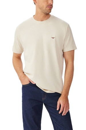 A man wears the RM Williams Parson T-Shirt, a light beige short sleeve top with a ribbed crew neck and small Longhorn embroidery on the chest, paired with dark blue jeans, standing against a plain white background.