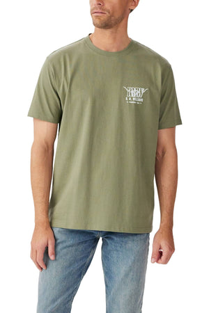 A man wears the RM Williams Prospect T-Shirt, a light olive green tee with a ribbed crew neck, short sleeves, and a small white logo on the left chest, paired with light blue jeans against a plain white background.