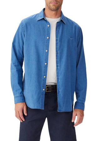 A man wears the RM Williams Arno Regular Indigo Shirt, a blue denim button-up with a French placket, over a white t-shirt, paired with black pants and belt. He stands facing forward, left hand by his side, right hand partly in his pocket.