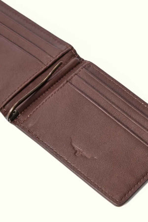 The RM Williams Farrier Bi Fold Wallet, crafted from premium brown leather, lies on a light surface. It features multiple card slots, a modern money clip, and a subtle embossed design on the lower part.