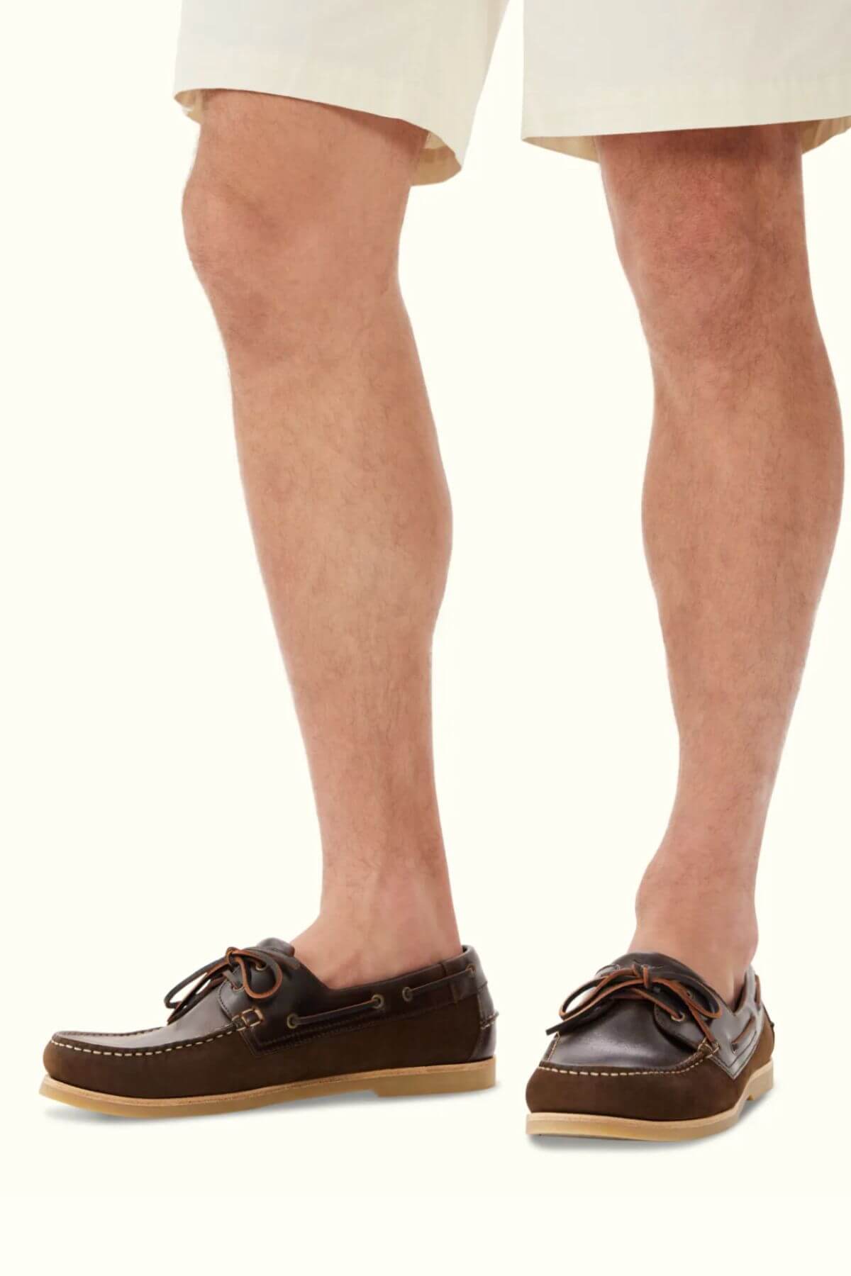 A person wearing beige shorts and RM Williams Hobart Boat Shoes in brown leather with tan soles and classic moccasin construction stands against a plain light background.