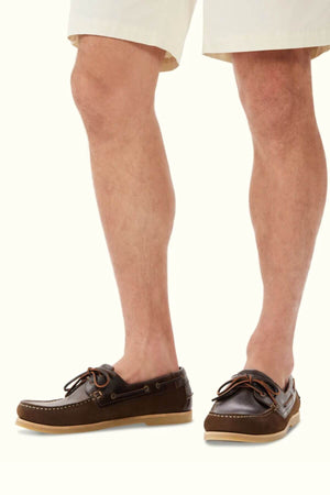 A person wearing beige shorts and RM Williams Hobart Boat Shoes in brown leather with tan soles and classic moccasin construction stands against a plain light background.