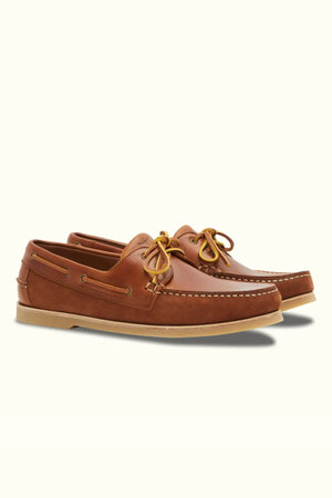 The RM Williams Hobart Boat Shoe features premium brown bovine leather with yellow laces, a moccasin toe, and tan rubber soles, elegantly displayed on a light background.
