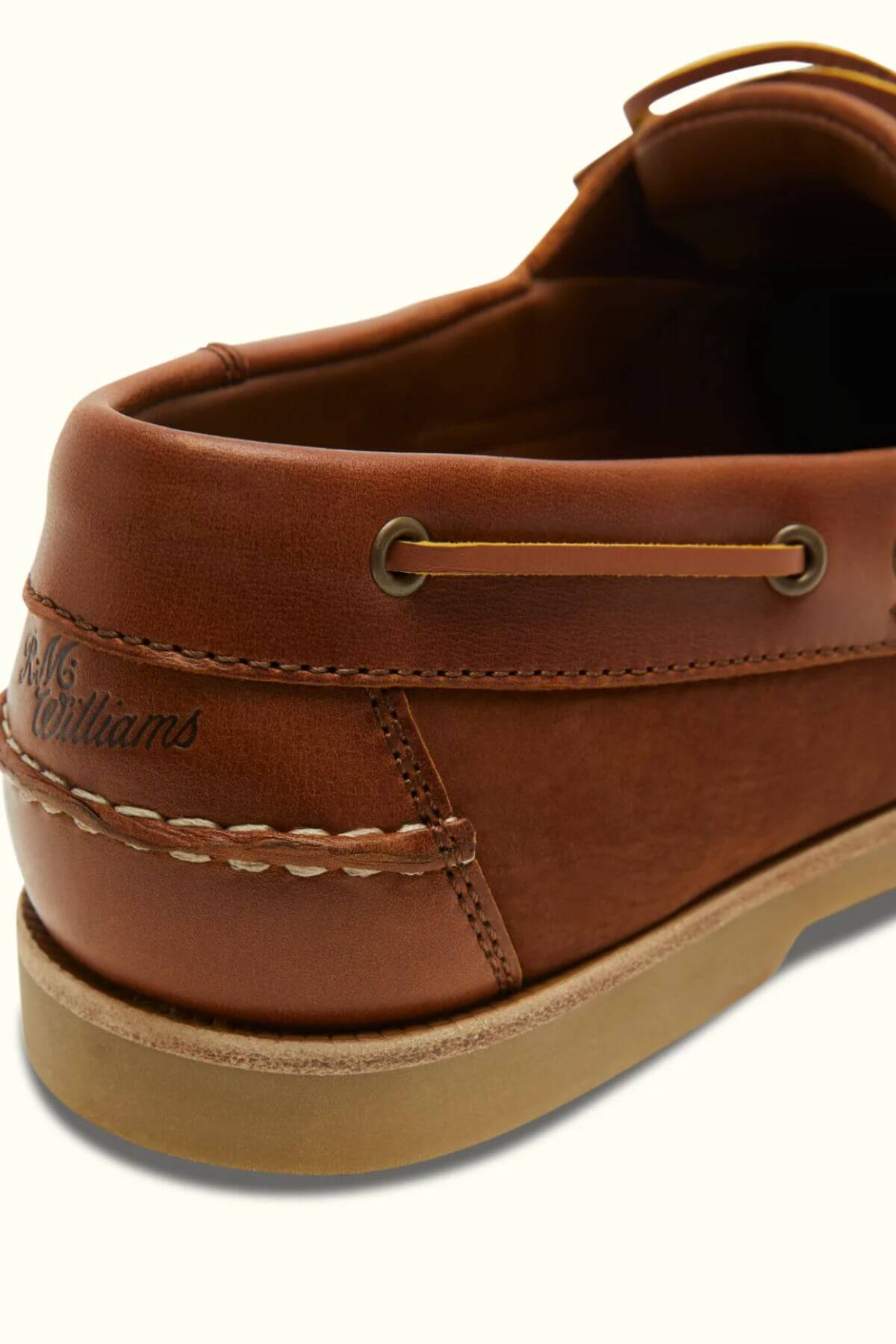 Close-up of a brown RM Williams Hobart Boat Shoe, crafted from bovine leather with the brand name engraved near the heel. The casual shoe showcases stitching details and metal eyelets, set on a light-colored sole.