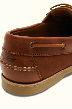 Close-up of a brown RM Williams Hobart Boat Shoe, crafted from bovine leather with the brand name engraved near the heel. The casual shoe showcases stitching details and metal eyelets, set on a light-colored sole.
