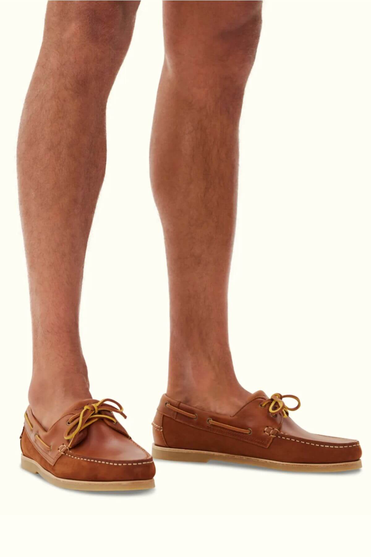 A person wearing RM Williams Hobart Boat Shoes, made from rich bovine leather with yellow laces, stands on a light floor. The shoes boast visible stitching and encapsulate timeless casual wear. The view captures the legs from the knees down.