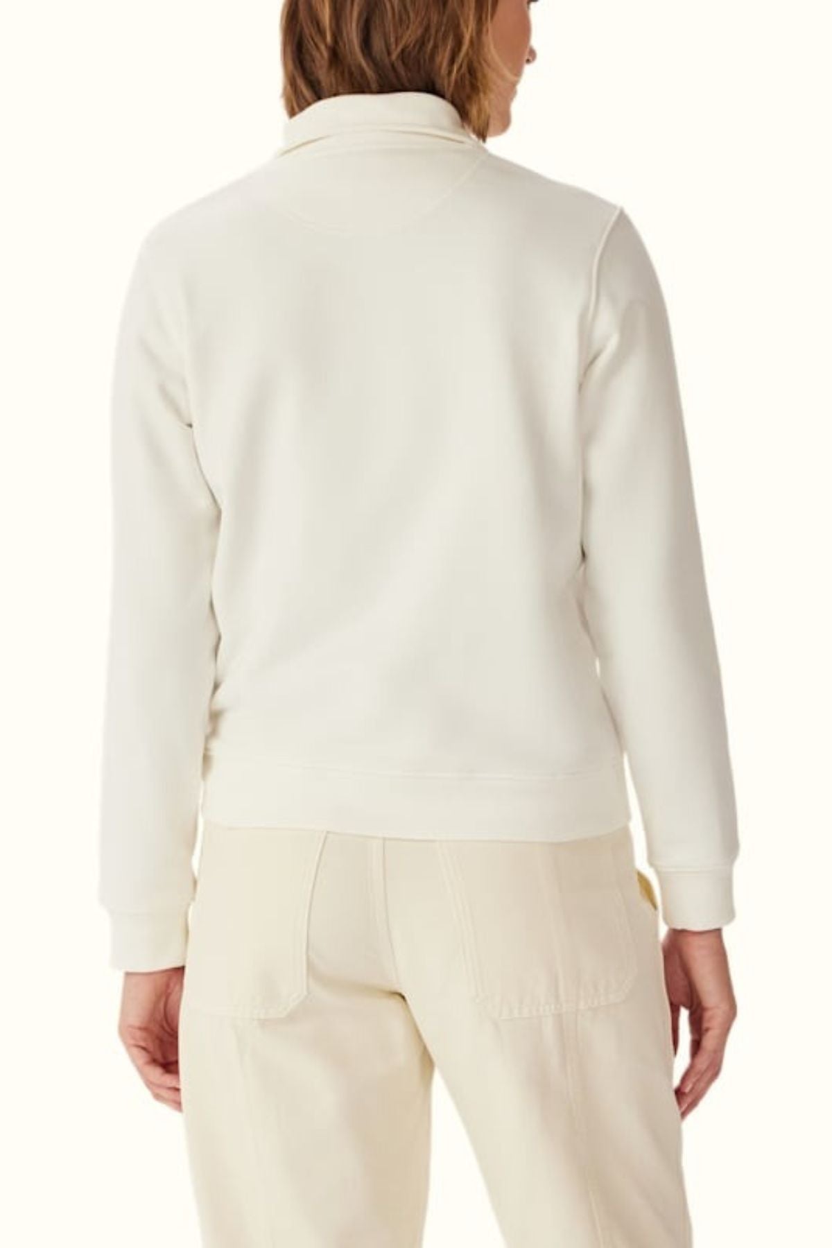 A person is shown from the back wearing the RM Williams Trickett Zip Neck Sweatshirt in cream with matching light pants, against a plain light background, their head turned slightly to one side.