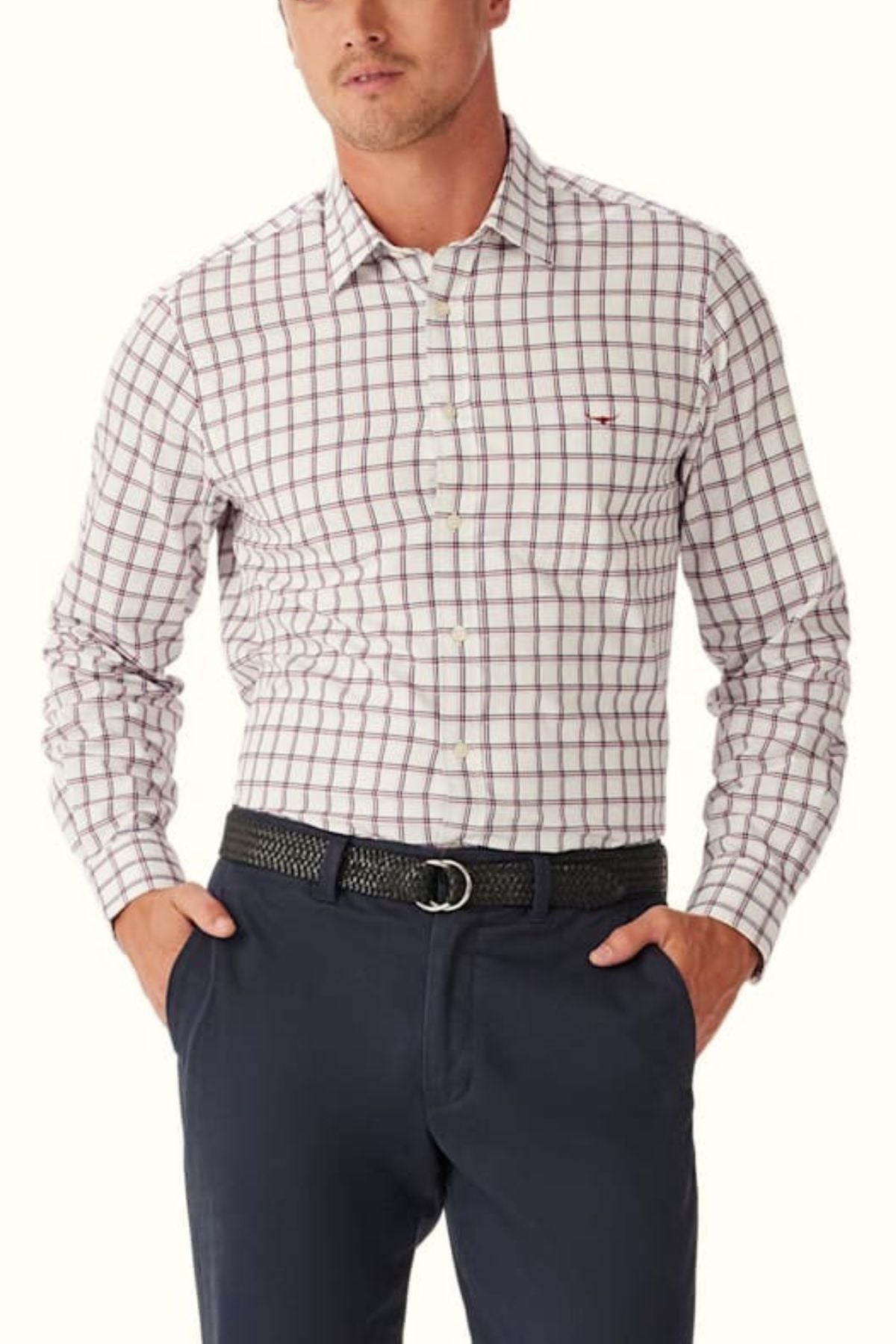 A man wears an RM Williams Classic Twill Check Shirt in white with red and blue checks, tucked into navy pants and secured with a black woven belt. Made from premium cotton, his hands rest casually in his pockets.