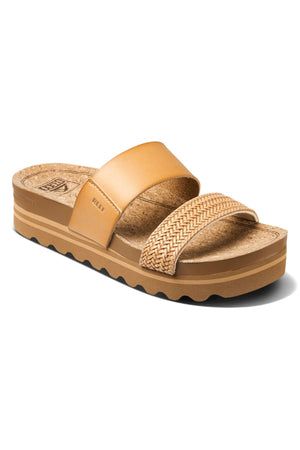 The REEF Cushion Vista Hi TX is a tan slide sandal with a cork footbed, smooth wide leather strap, braided toe strap, and a thick, slightly raised sole with wavy black tread for comfort.