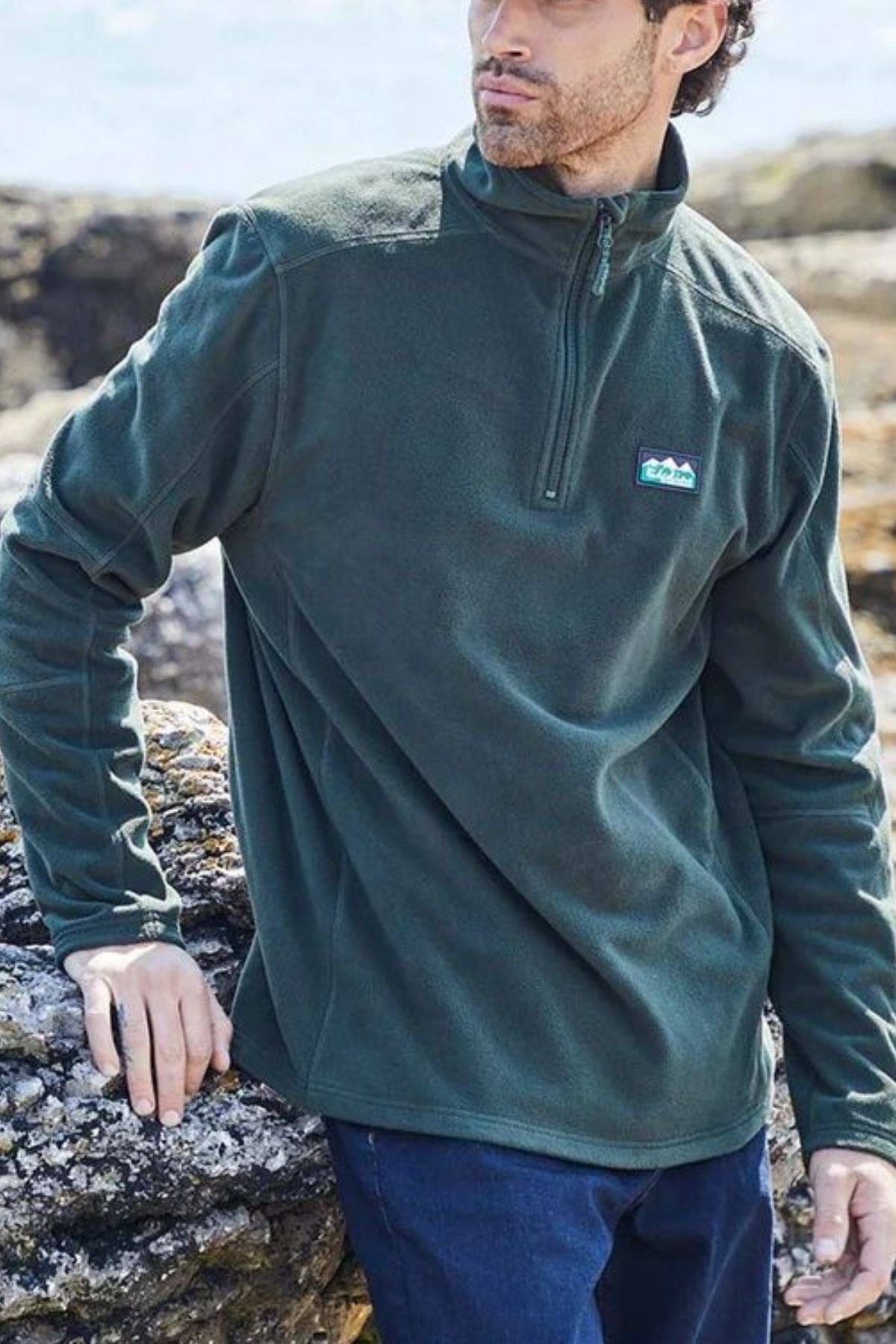 A man stands on rocky terrain outdoors, looking to the side, wearing the Ridgeline Narvik Fleece—a dark green lightweight zip-neck pullover with a small patch logo.