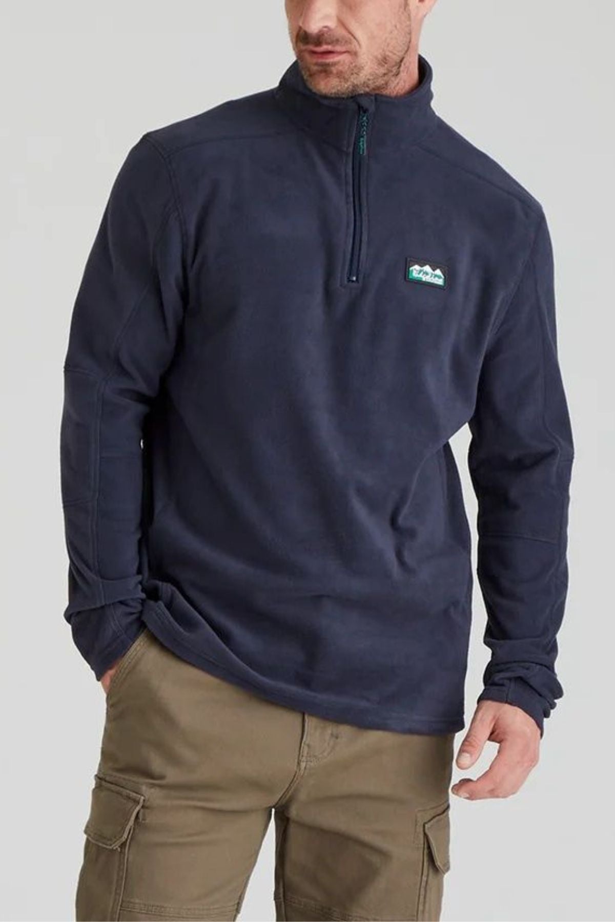 A man wears the Ridgeline Narvik Fleece quarter-zip in navy blue, featuring a small chest patch, paired with tan cargo pants. He stands against a light background, embodying outdoor performance style.
