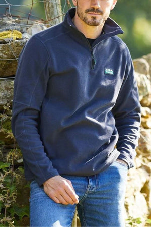 A man with a short beard wears a navy blue Ridgeline Narvik Fleece zip-up and blue jeans, sitting outdoors by a stone wall with greenery—ideal for outdoor performance.
