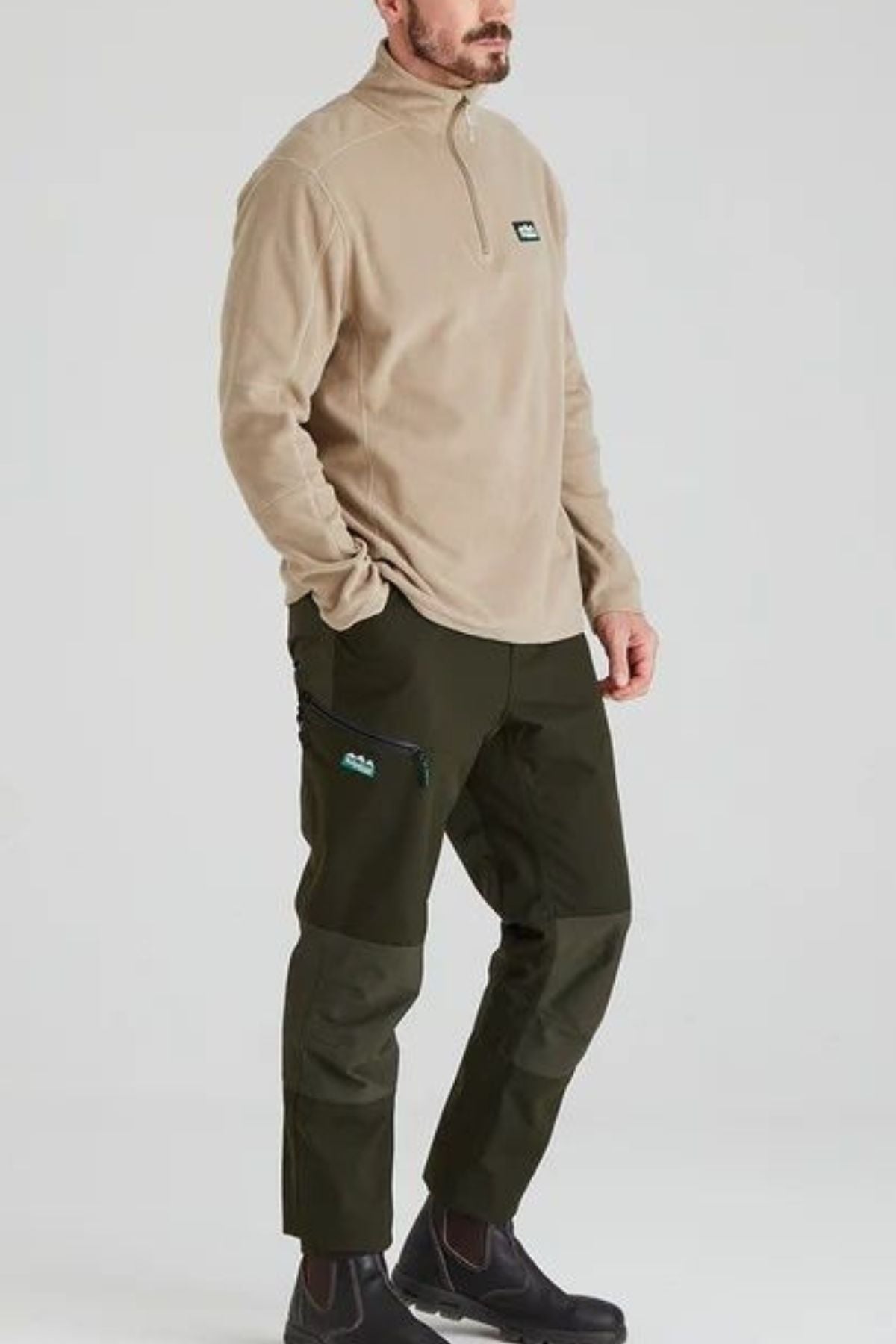 A man wearing a Ridgeline Narvik Fleece zip-neck pullover, dark green cargo pants with zipper details, and black boots stands against a plain light gray background.