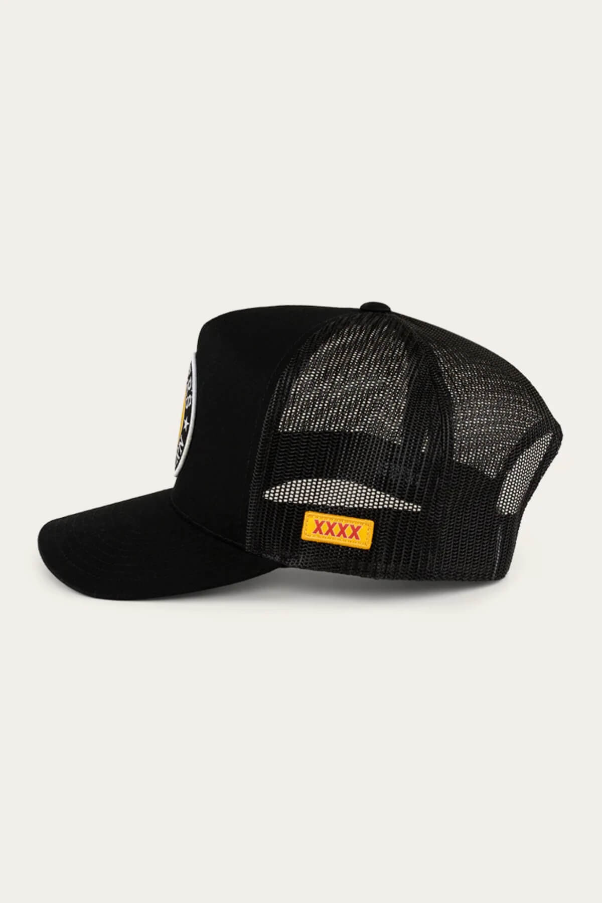 The Ringers Western Can Trucker is a black cap with a twill front and poly mesh back, featuring a round patch with black, yellow, and white embroidery on the front panel, and a yellow rectangular patch with red "XXXX" text on the side against a light gray background.