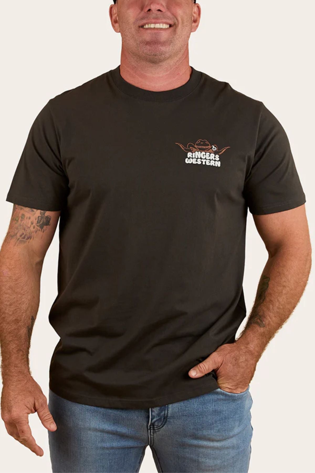 A smiling man with forearm tattoos wears the Ringers Western Country Club Loose Fit T-Shirt in black, featuring text and a small left chest graphic, paired with blue jeans against a plain background.