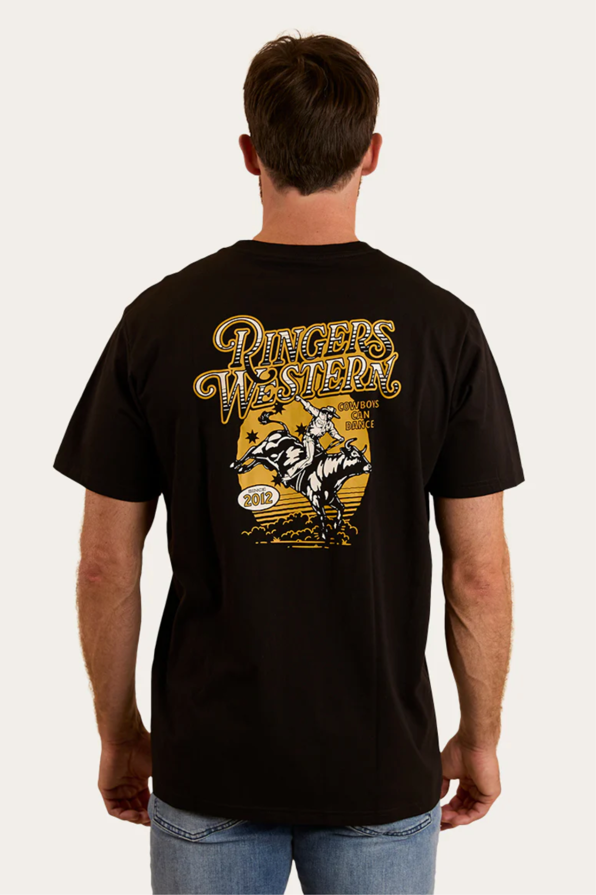 A man wears the Ringers Western Cowboys Can Dance Loose Fit T-Shirt, featuring a yellow and white "Cowboys Can Dance" graphic, a cowboy riding a bull, stars, and the year 2012 on the back.