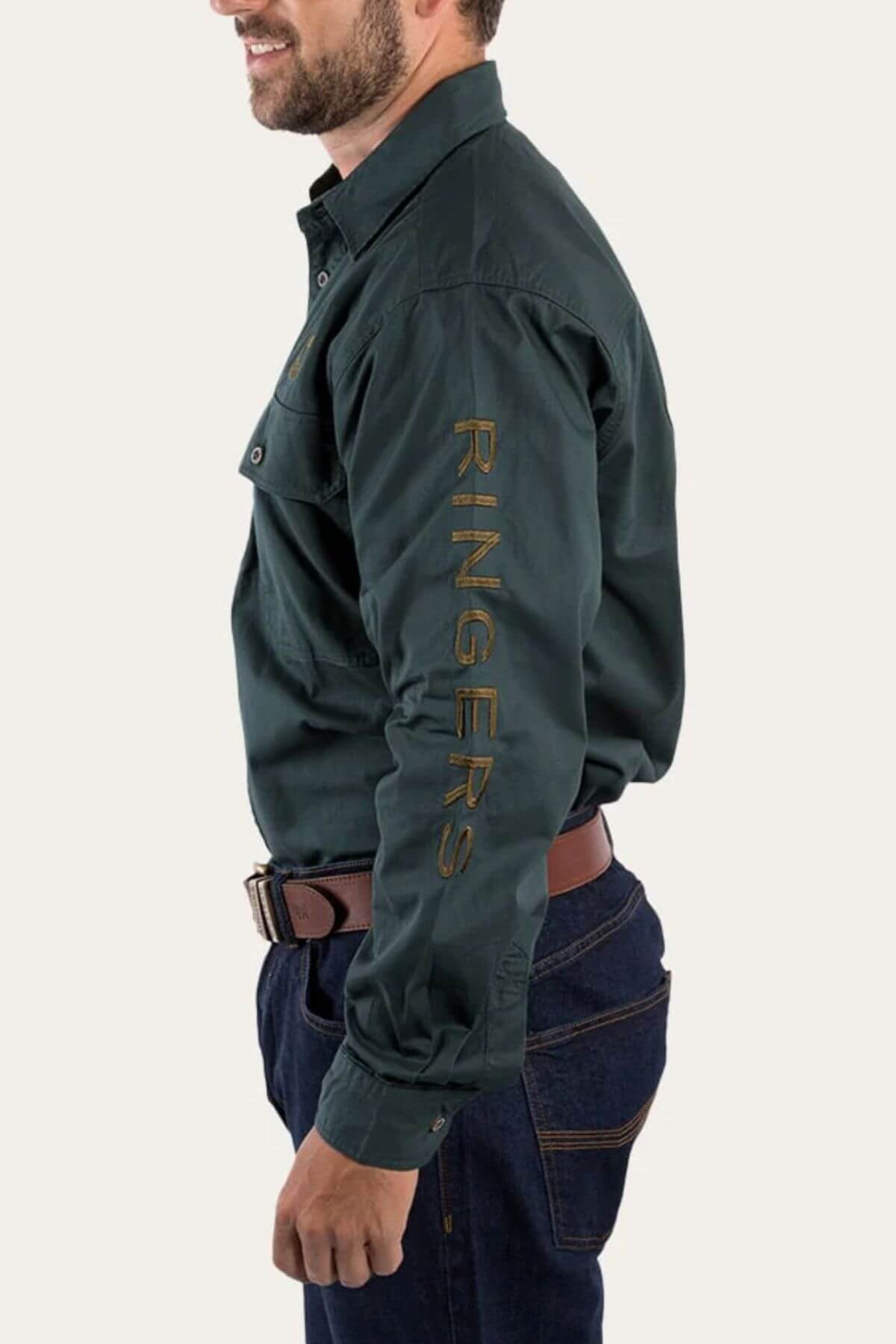 Wearing the Ringers Western Hawkeye Embroidered Shirt in dark green premium cotton with "RINGERS" on the sleeve, a person stands with their right side facing forward. They match it with dark jeans and a brown belt against a plain white background.