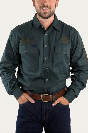 A man in a dark green Ringers Western Hawkeye Embroidered Shirt, showcasing patches and embroidery, long sleeves, and two chest pockets. Paired with dark jeans and a brown belt with a silver buckle, his hands rest near his waist, exuding casual style.
