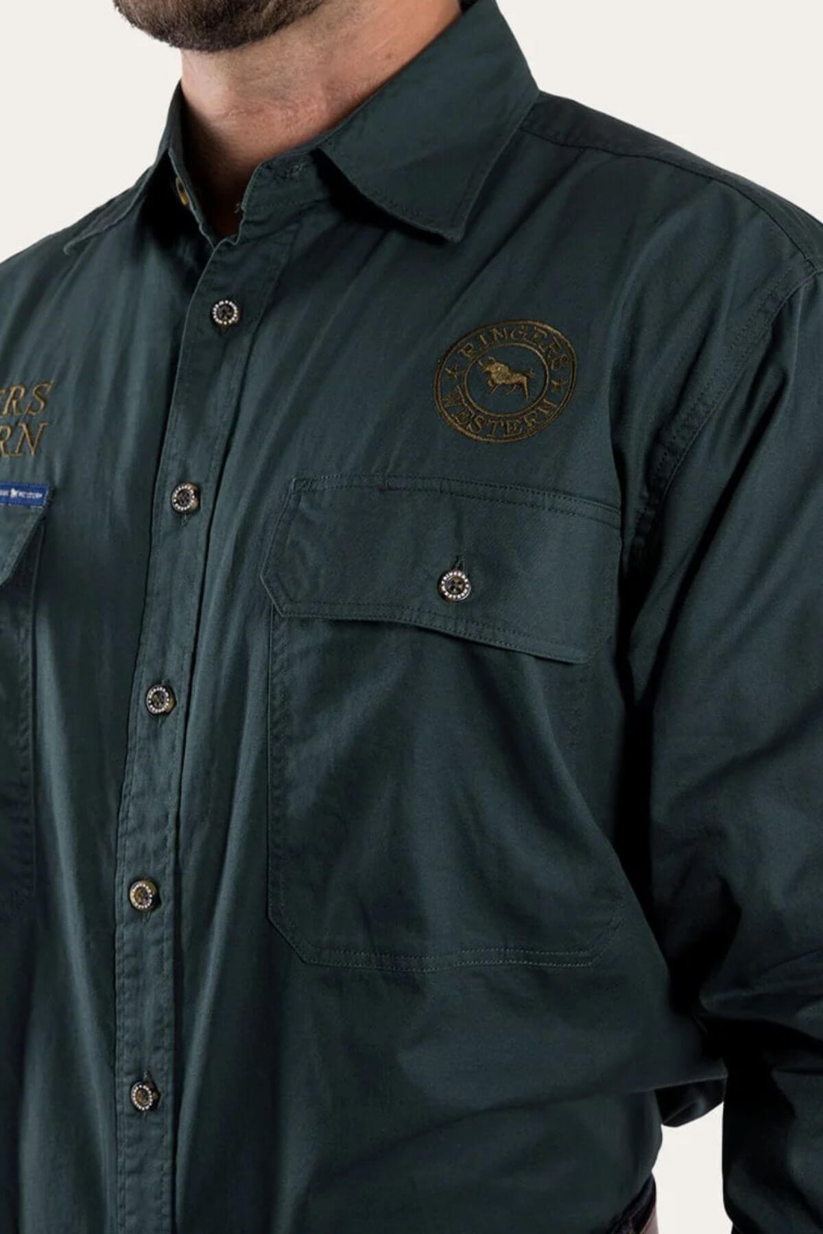 A person wears a Ringers Western Hawkeye Embroidered Shirt in dark green, made of premium cotton with an emblem and text patch on the chest. It features button-up style, visible stitching, and buttoned chest pockets; their face isn't visible.