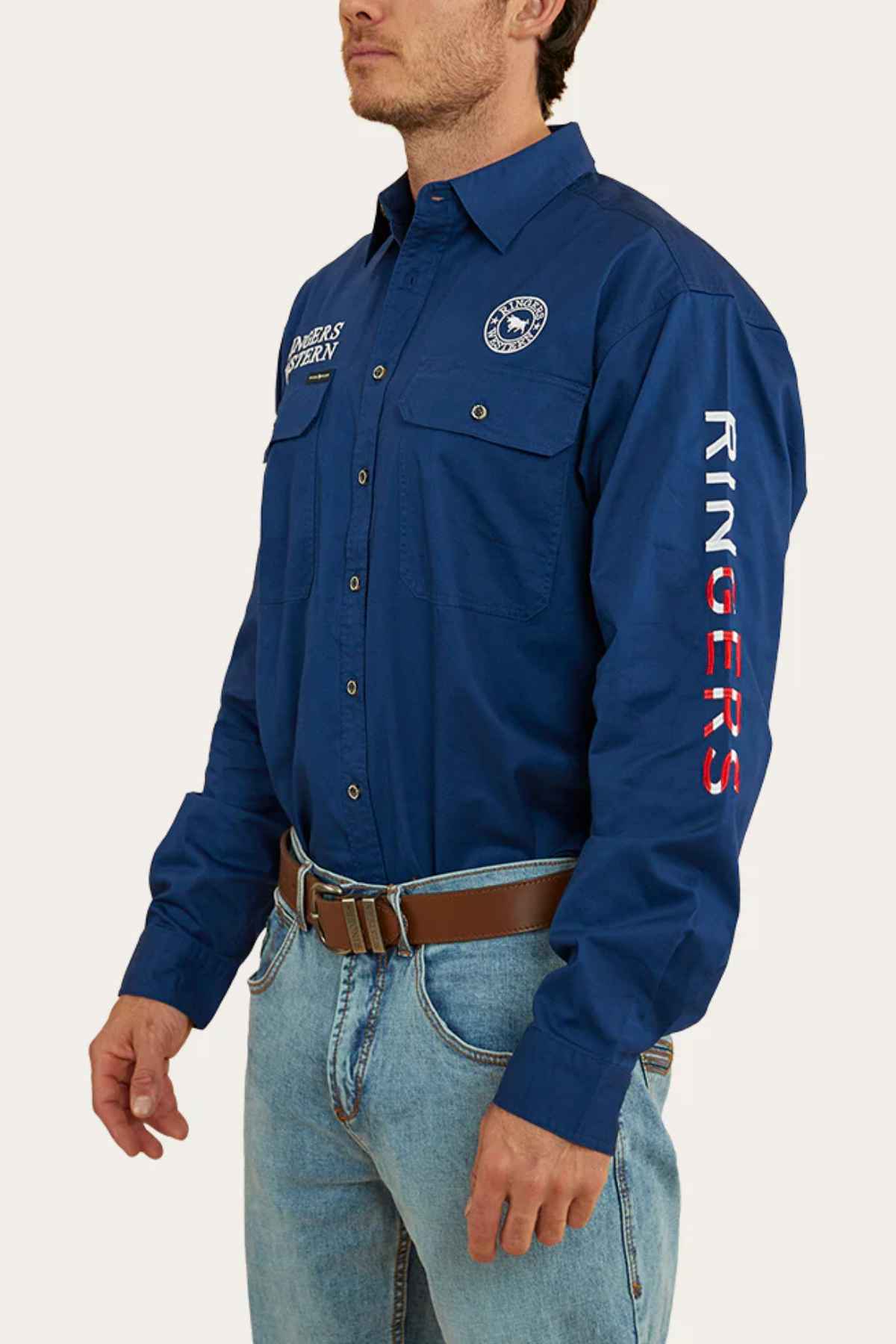 A person is sporting a Ringers Western Hawkeye Flag Embroidered Shirt, adorned with patches and "RINGERS" on the sleeve. They pair it effortlessly with light blue jeans and a brown belt, capturing a laid-back yet stylish vibe.