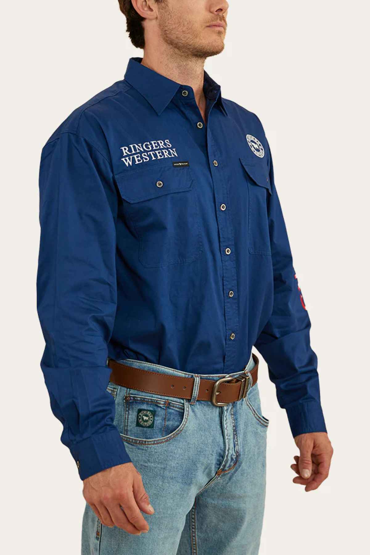 A man is wearing the blue Ringers Western Hawkeye Flag Embroidered Shirt with long sleeves and button-up style, which features embroidered "Ringers Western" logos. He pairs it with light blue jeans and a brown belt, creating a rugged yet polished appearance.