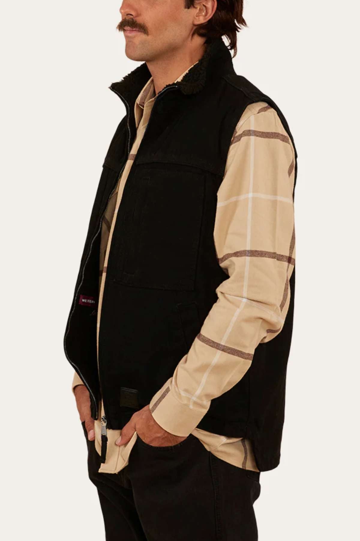 A man with a moustache wearing a Ringers Western High Country Vest made of durable cotton canvas in black, layered over a beige plaid shirt, stands with his hands in his pockets against a plain white background.